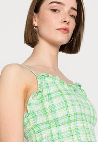 Vestido de verão em xadrez verde e branco com alças com babados, corpete justo e tecido texturizado. Os detalhes incluem pequenos toques de amarelo claro no padrão.
