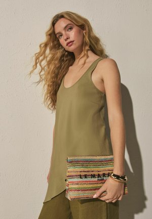 Femeie cu păr lung, ondulat și blond purtând un top kaki fără mâneci și ținând o geantă tip plic țesută multicolor cu text.