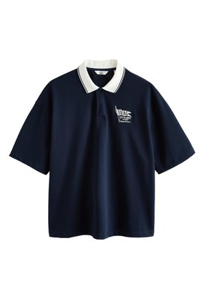 Junge Person trägt einen beigen Bucket Hat, ein navyfarbenes Oversized-Poloshirt, grüne karierte Shorts, weiße Socken und beige Slip-On-Schuhe und steht vor einer schlichten Wand.