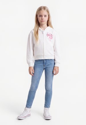 Jeune fille aux longs cheveux blonds se tient face à l'avant, portant un sweat à capuche blanc zippé avec un motif rose, un jean bleu clair et des baskets blanches montantes.