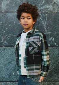 Jongen met krullend haar, gekleed in een groen en zwart geruit shirt over een wit T-shirt met opdruk, staande tegen een donkere groene marmeren muur.
