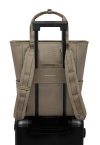 Beige tote bag met dubbele schouderbanden, geplaatst op de handgreep van een zwarte koffer. Heeft een gladde textuur en minimale merkidentificatie.