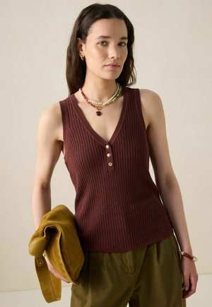 Vrouw met lang donker haar, gekleed in een bruin mouwloos gebreid topje, olijfgroene broek, met een kralenketting, armband en een mosterdkleurige suède tas.