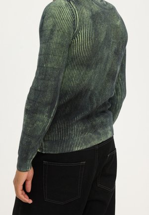 Man wearing a green and black ribbed long-sleeve sweater with a subtle gradient, paired with black pants with visible back pockets.