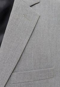 Blazer grigio con un design strutturato, realizzato in tessuto texturizzato. Presenta un colletto a scollo a V, una tasca sul petto sinistro e una tasca laterale.