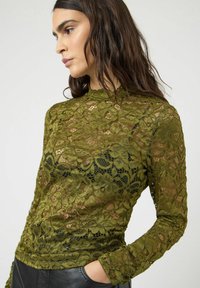 Oliwkowo-zielony koronkowy top z misternymi wzorami kwiatowymi, wysokim kołnierzem i długimi rękawami; cienka, półprzezroczysta tkanina na czarnym podkładzie.
