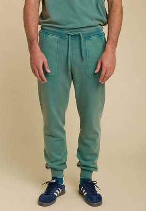 Jogging en coton vert avec taille élastique, cordon de serrage et deux poches avant. Associé à des baskets bleues avec des rayures blanches.