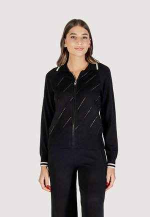 Žena oblečená v čiernom svetri na zips s diagonálnymi vzormi a zodpovedajúcimi čiernymi nohavicami, stojaca pred jednoduchým svetlým pozadím.