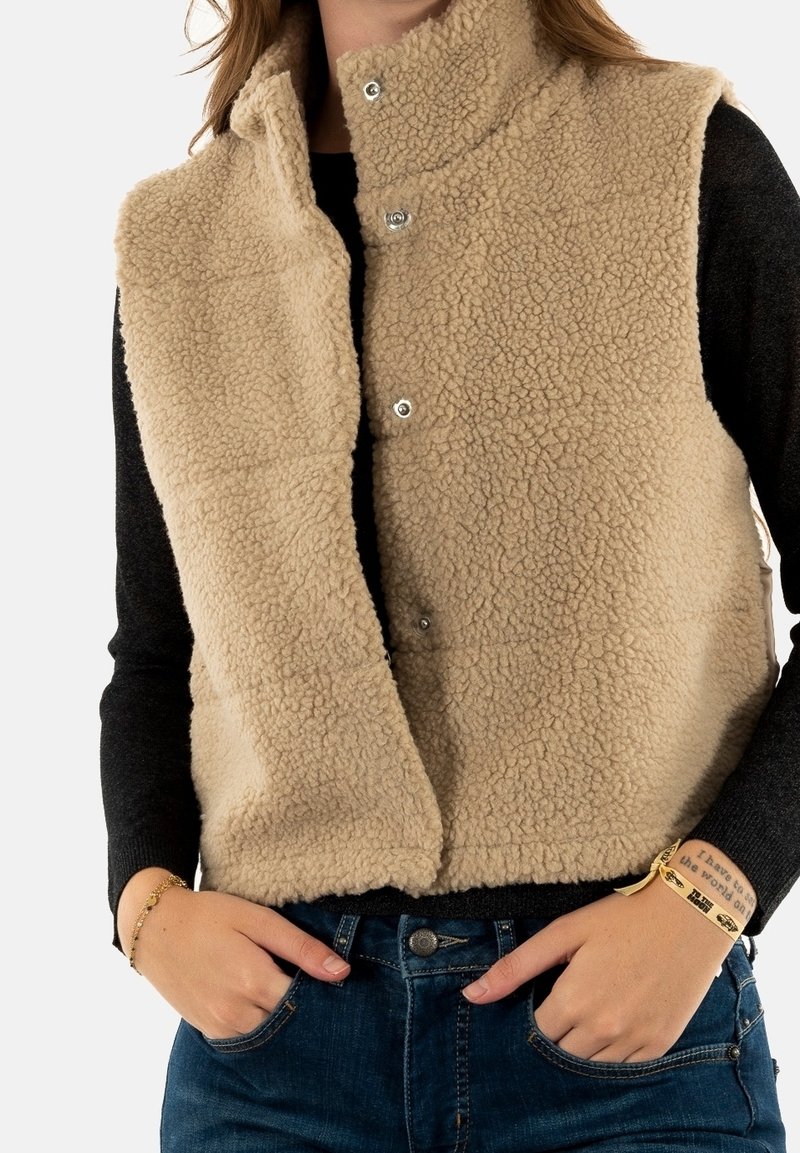Personne portant un gilet beige duveteux sur une chemise noire à manches longues, mains dans les poches d’un jean bleu, bracelets visibles aux deux poignets.