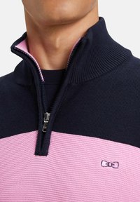 Sweatshirt zippé bleu marine et rose avec un col côtelé. Présente une rayure rose texturée et un petit logo sur la section rose.