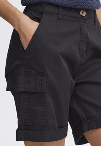 Handen i fickan på svarta cargoshorts med knapp och uppviktig fåll, bärs tillsammans med en mörkblå stickad topp.