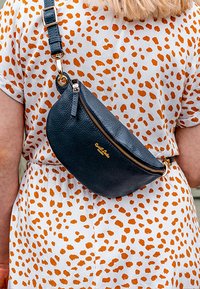 Bolso cruzado de cuero negro con forma curvada, superficie texturizada, cremallera dorada y logo en la parte frontal, llevado sobre un vestido blanco con manchas naranjas.
