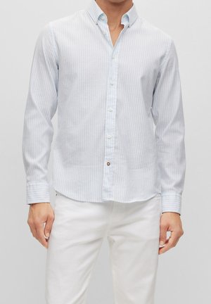 Mann trägt hellblau-weiß gestreiftes langärmliges Hemd mit weißen Hosen vor schlichtem grauem Hintergrund.