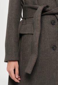 Brown wool coat with a tie belt, featuring a textured finish, two buttons, and a front pocket on the left side.
