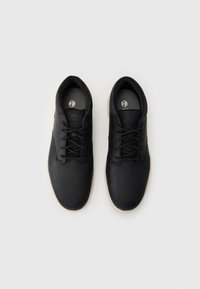 Zapatillas de cuero negro con cordones planos y puntera redonda. Presentan un logo en la lengüeta y una suela de goma con una capa inferior de color claro.