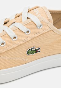 Sneaker in tela beige con lacci bianchi, suola bianca e logo di coccodrillo verde ricamato sul lato.