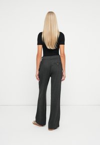 Persoon met lang blond haar staat met de rug naar ons toe, draagt een zwart T-shirt met korte mouwen, grijze high-waisted wijde broek en casual schoenen.
