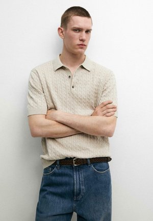 Jonge man met kort haar staat met armen over elkaar gekruist, draagt een beige kabelgebreide polo en blauwe spijkerbroek tegen een effen witte achtergrond.