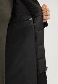 Hand die een open zwarte gewatteerde jas vasthoudt en een binnenzak met rits toont tegen een achtergrond van donkere kleding.