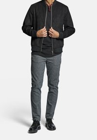 Černá suede bomber bunda s žebrovaným lemem, nošená přes černou košili, spárována se strukturovanými šedými kalhotami a černými šněrovacími botami.
