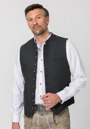 Mann trägt traditionelle bayerische Tracht mit weißem Hemd, dunkler Knopfwest und bestickter beiger Lederhose.