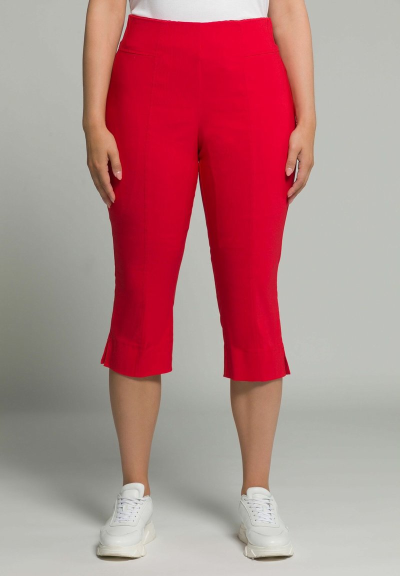 Pantalons capri rouges en tissu léger, avec un design ajusté, des coutures latérales et un ourlet évasé, associés à des baskets blanches.