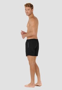 Shorts de bain noirs avec taille élastique, présentant des accents latéraux bleus. Le modèle se tient pieds nus dans une position neutre.