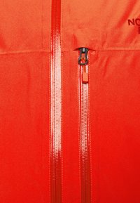 Tela de chaqueta naranja brillante con dos cremalleras impermeables verticales, una cremallera de bolsillo más pequeña parcialmente abierta y texto parcial "THE NORTH FACE."