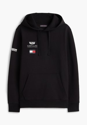 Νέος άνδρας που στέκεται με τα χέρια εν μέρει στις τσέπες, φορώντας μια μαύρη μπλούζα με κουκούλα με λογότυπα, μπεζ παντελόνια και άσπρα παπούτσια σε λευκό φόντο.