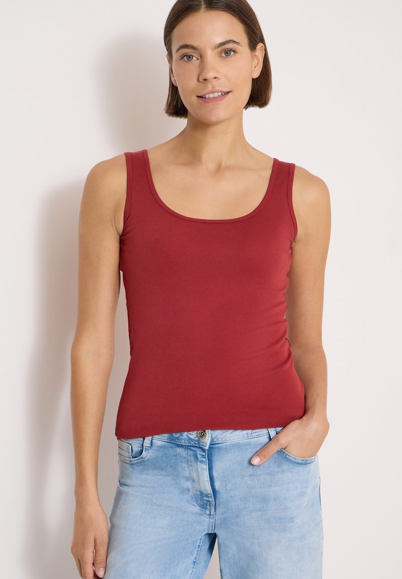 Femme aux cheveux courts et bruns coiffée d'un débardeur rouge sans manches et d'un jean bleu clair, debout avec une main dans sa poche.