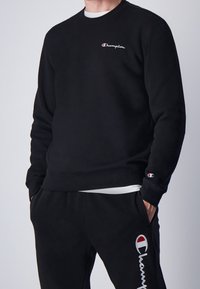 Sudadera negra con cuello redondo y logotipo bordado de "Champion". Los pantalones de chándal cuentan con un logotipo a juego y bolsillos laterales. Tejido suave.