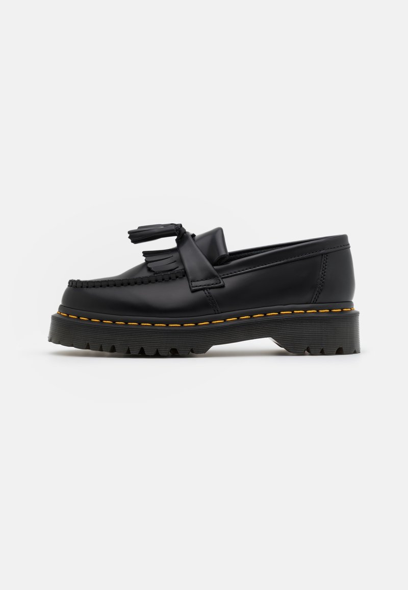 Mocassin en cuir noir avec semelle épaisse, coutures jaunes, détail pampille et semelle extérieure rainurée sur fond blanc.