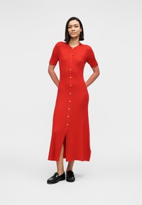 Κόκκινο πλεκτό φόρεμα μήκους μέχρι τα γόνατα με κοντά μανίκια, στρογγυλή λαιμόκοψη, κουμπιά μπροστά και πλαϊνή ανοίγματα, φορεμένο με μαύρα παπούτσια.