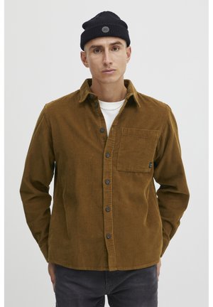 Bruine corduroy overhemd met knoopsluiting, een borstzak en een gestructureerde afwerking. Draag het over een wit shirt, in combinatie met een zwarte beanie.