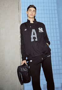Sort træningsbeklædning med en zip-up jakke med New York Yankees-logo, et mønster med "A" på skjorten og matchende sweatpants. En sort taske bæres.