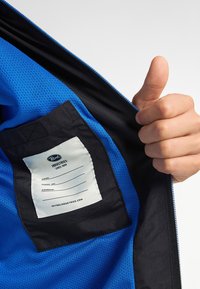 Căptușeală din plasă de poliester albastru cu accente negre. Include o etichetă cu logo-ul "Petrol Industries" și zone pentru informații personale. Închidere cu fermoar.