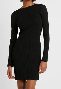 Svart ribbad långärmad klänning med en figursydd silhuett. Texturerat tyg, rund halsringning och knälång design lyfter fram dess eleganta utseende.