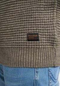 Gehaakte, grijze trui met ribgebreide zoom, voorzien van een oranje en zwarte geweven label. De achtergrond bevat lichtblauwe spijkerbroeken.