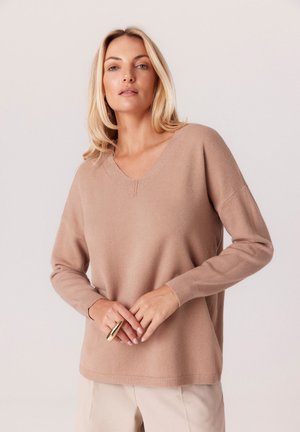 Jasnobrązowy sweter w serek, wykonany z miękkiego materiału, z długimi rękawami i prążkowanymi mankietami, stylizowany na beżowych spodniach z złotym pierścionkiem.