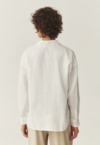 Persona con cabello rizado, vistiendo una camisa de lino blanca de manga larga y pantalones beige, vista desde atrás sobre un fondo liso.