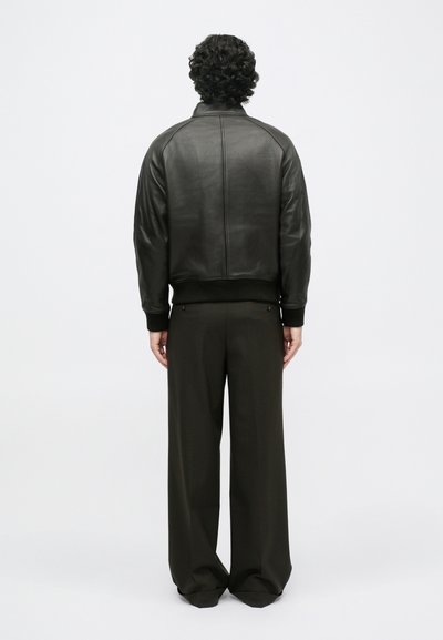 Μαύρη δερμάτινη τζάκετ bomber με ριγωτό τελείωμα και μανίκια, χαρακτηρισμένη από λεία υφή και ελαφρώς υψωμένο γιακά. Συνδυάζεται με φαρδιά, σκούρα παντελόνια.