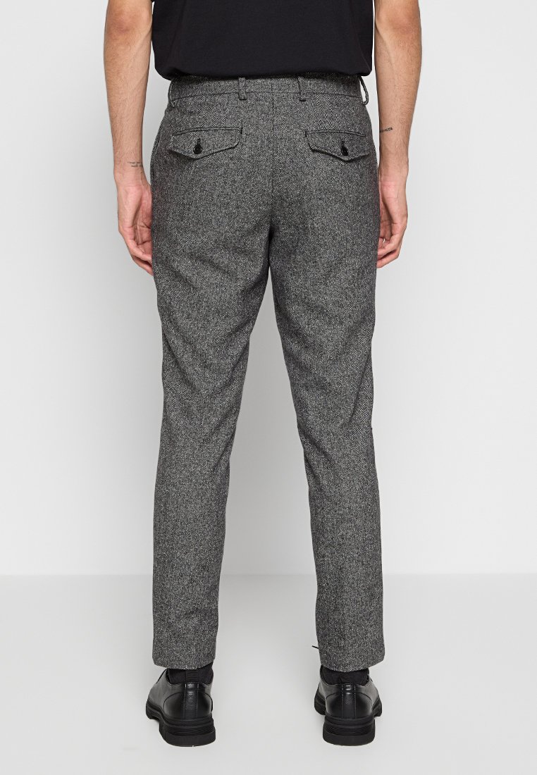 Uomo che indossa pantaloni grigi su misura con motivi, tasche posteriori con bottoni, calzini neri, scarpe eleganti nere e una camicia nera, in piedi di spalle.