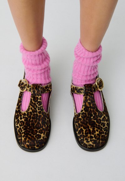 Des jambes portant des chaussettes côtelées rose vif, associées à des chaussures Mary Jane à imprimé léopard avec des boucles dorées, sur un fond clair uniforme.