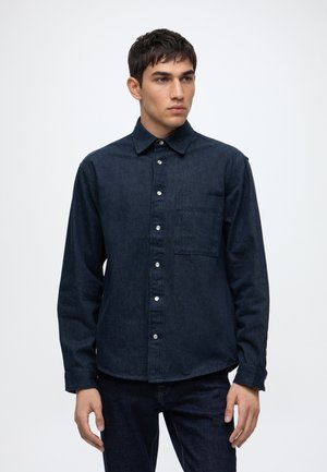 Jeune homme portant une chemise en jean bleu foncé à boutons avec une poche poitrine, debout devant un fond clair uni.