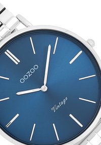 Montre en argent avec un cadran bleu mat, index et aiguilles argentés. La marque "oozoo" et "Vintage" sont affichées sur le cadran. Bracelet en métal.