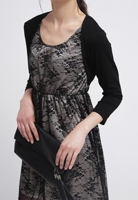 Musta kardigan hopea- ja mustalace-mekon päällä, jossa on istuva vyötärö. Asusteeseen kuuluu musta clutch-laukku, jota pidetään oikeassa kädessä.