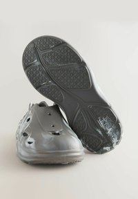 Grå gummiklippere med en glat overflade, ventilationshuller og teksturerede såler. Let hævet hæl, designet til komfort og nem påklædning.