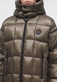 Personne portant une doudoune brillante vert olive avec col haut, fermeture éclair noire, deux poches zippées et un patch logo rond noir sur la poitrine.