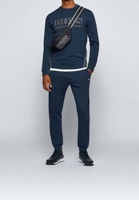 Felpa blu navy con stampa del logo, abbinata a pantaloni jogger con taglio affusolato e sneakers nere, completata da una borsa a tracolla. Texture liscia, design casual.