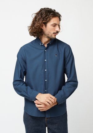 Homme aux cheveux bouclés mi-longs portant une chemise bleu marine boutonnée avec un petit logo d'arbre sur la poitrine, debout les mains jointes.